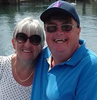 Norrms and his wife are smiling at the camera wearing sun glasses in front large body of water.