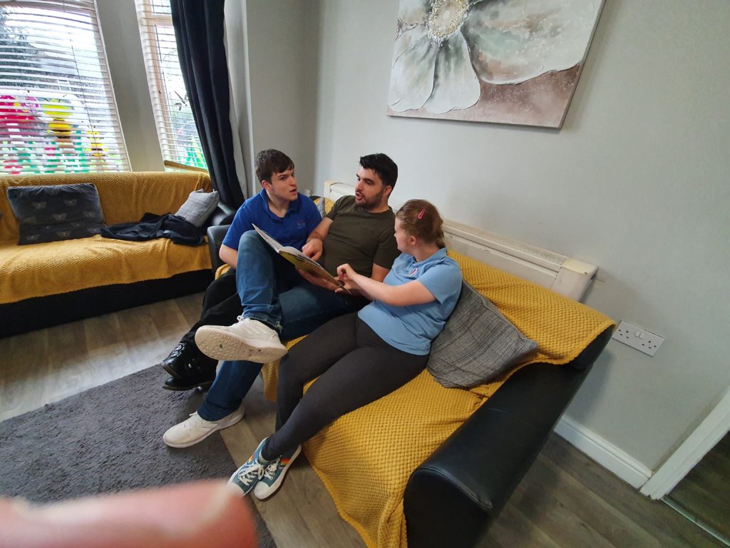 Alexander sat on the sofa reading with two young people he supports