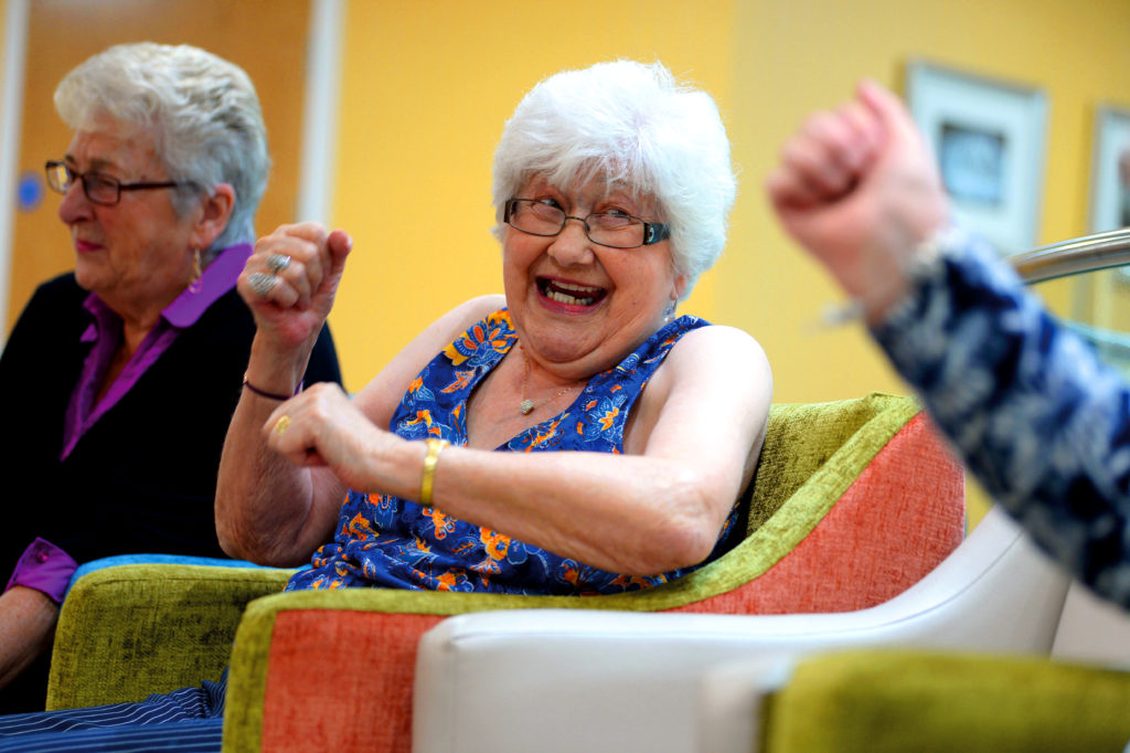 An older lady with a wide smile is looking to her friend doing a work out in her chair