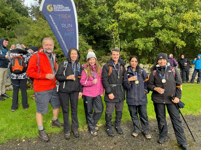 Bevan Brittan team stand in a line smiling at the camera in hiking gear, standing in front of a large tree and hiking companies banner.