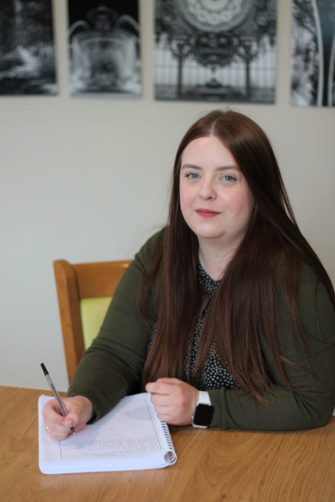 Chloe sits at a desk, smiling at the camera writing with a pen and paper