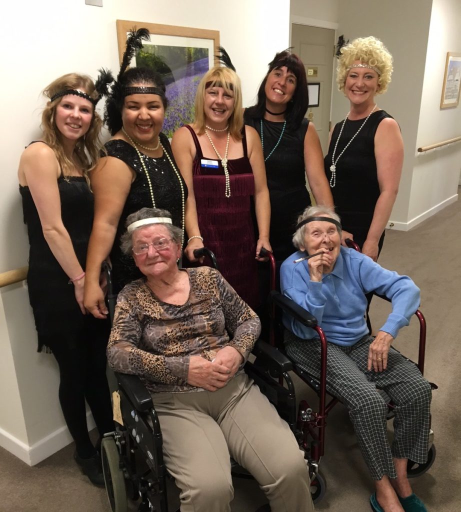 A group of female care workers dressed in 20's outfits with two female residents in wheel chairs. 