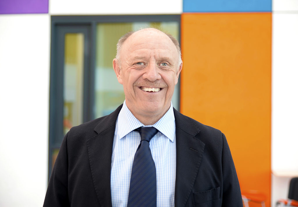 Time looks smiling into the camera in a suit and time, the building behind has several blocks of colour. 