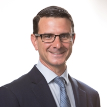 Vincent smiles to the camera in a suit with a blue tie, smiling in glasses at the camera