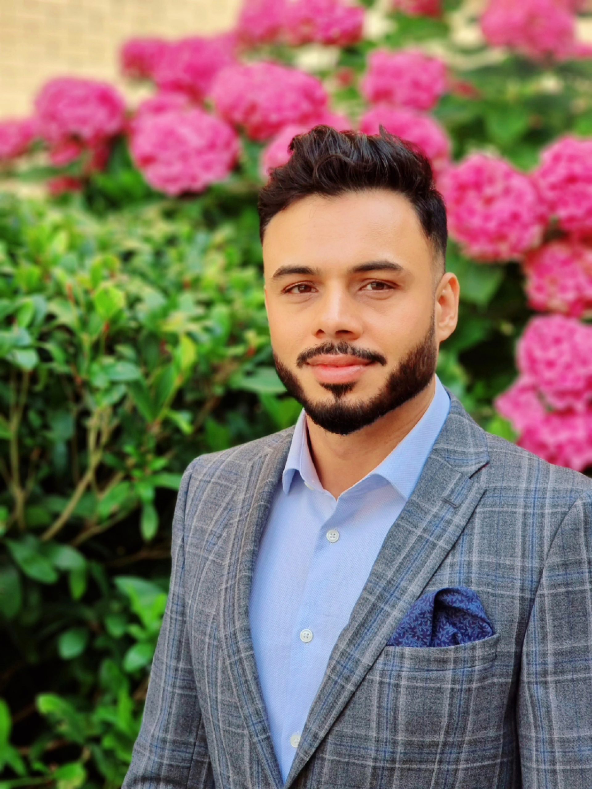 Zahid stands in a grey suit in front of a green bush with pink flowers