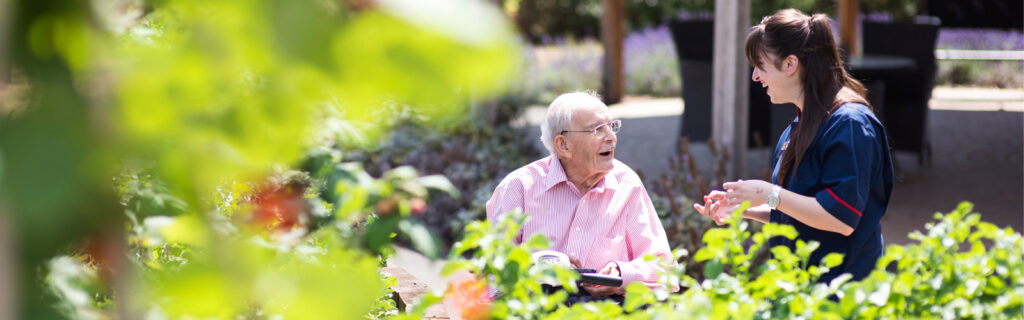 an older man and his career stand behind a tree talking. He is sat down and she is explaining something to him. 