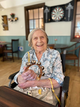 Older lady is sat at a table in a care home day room, holding a small hand instrument with bells, smiling at the camera. 