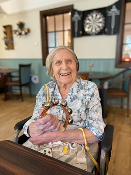 Older lady is sat at a table in a care home day room, holding a small hand instrument with bells, smiling at the camera.